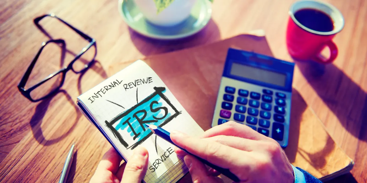 Hands sketching the letters IRS on a notepad beside a calculator and glasses on a wooden desk